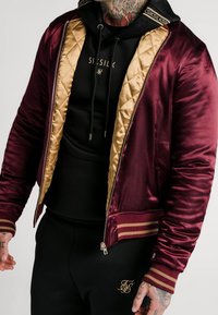 Mørkerød satin bomberjakke med en guld quiltet foring, ribbede burgunder og guld manchetter samt lynlåslukning. Sort hættetrøje under har et logo.