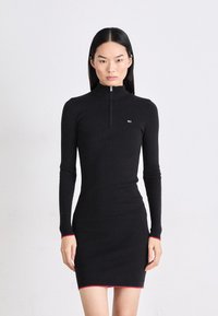 Rochie neagră cu nervuri, cu guler înalt și fermoar, având mâneci lungi și accente roșii la manșete și la tiv.