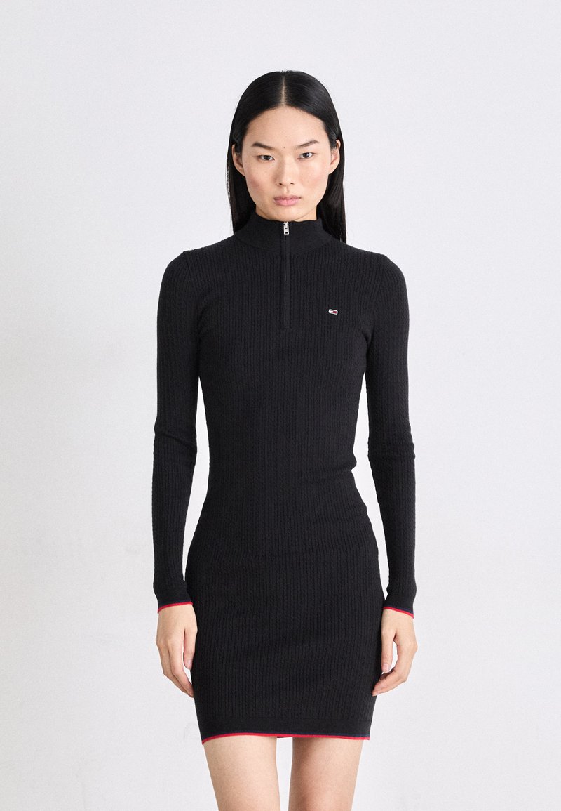 Rochie neagră cu nervuri, cu guler înalt și fermoar, având mâneci lungi și accente roșii la manșete și la tiv.