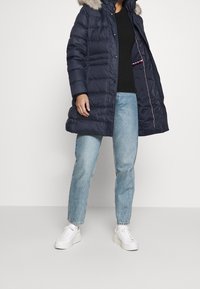 Námořnický puffer kabát s kapucí lemovanou kožešinou, se sklapovacími knoflíky, prošívaným designem a kontrastními detaily na vnitřní kapse. Nosený s světlými džínami a bílými teniskami.