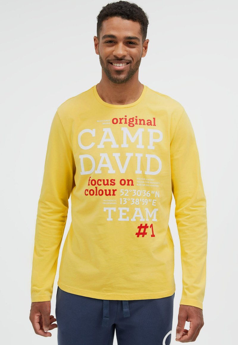 Lächelnder Mann, der ein gelbes Langarmshirt mit rotem und weißem Text sowie marineblaue Hose trägt, vor einem schlichten weißen Hintergrund.