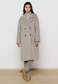 Beige dubbelzijdige wollen jas met grote zwarte knopen, brede revers en een losse pasvorm, gecombineerd met een grijze turtleneck en lichtgekleurde broek.