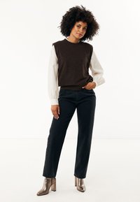 Maglione senza maniche marrone sopra una camicia a maniche lunghe bianca con texture, abbinato a jeans neri e stivaletti con tacco metallici.