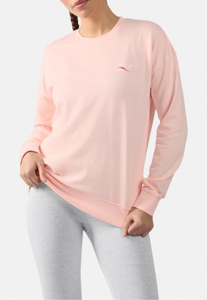 Vrouw draagt een lichtroze langemouwen Reebok-shirt en lichtgrijze leggings, terwijl ze de zoom van het shirt zachtjes met beide handen naar beneden trekt.