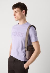 T-shirt en coton lavande avec un logo "BOSS" audacieux, associé à un pantalon noir et un sac à épaule texturé marron avec une fermeture éclair et un détail de logo.