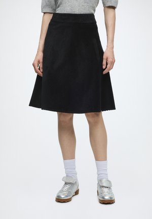 Personne portant une jupe trapèze en velours côtelé noire, un haut gris, des chaussettes blanches montantes et des chaussures argentées métalliques à lacets avec une bride.