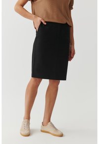 Čierna sukňa po kolená s elegantným dizajnom, vyrobená z hladkej tkaniny. Obsahuje bočné vrecká a je skombinovaná so svetlohnedými neformálnymi teniskami.