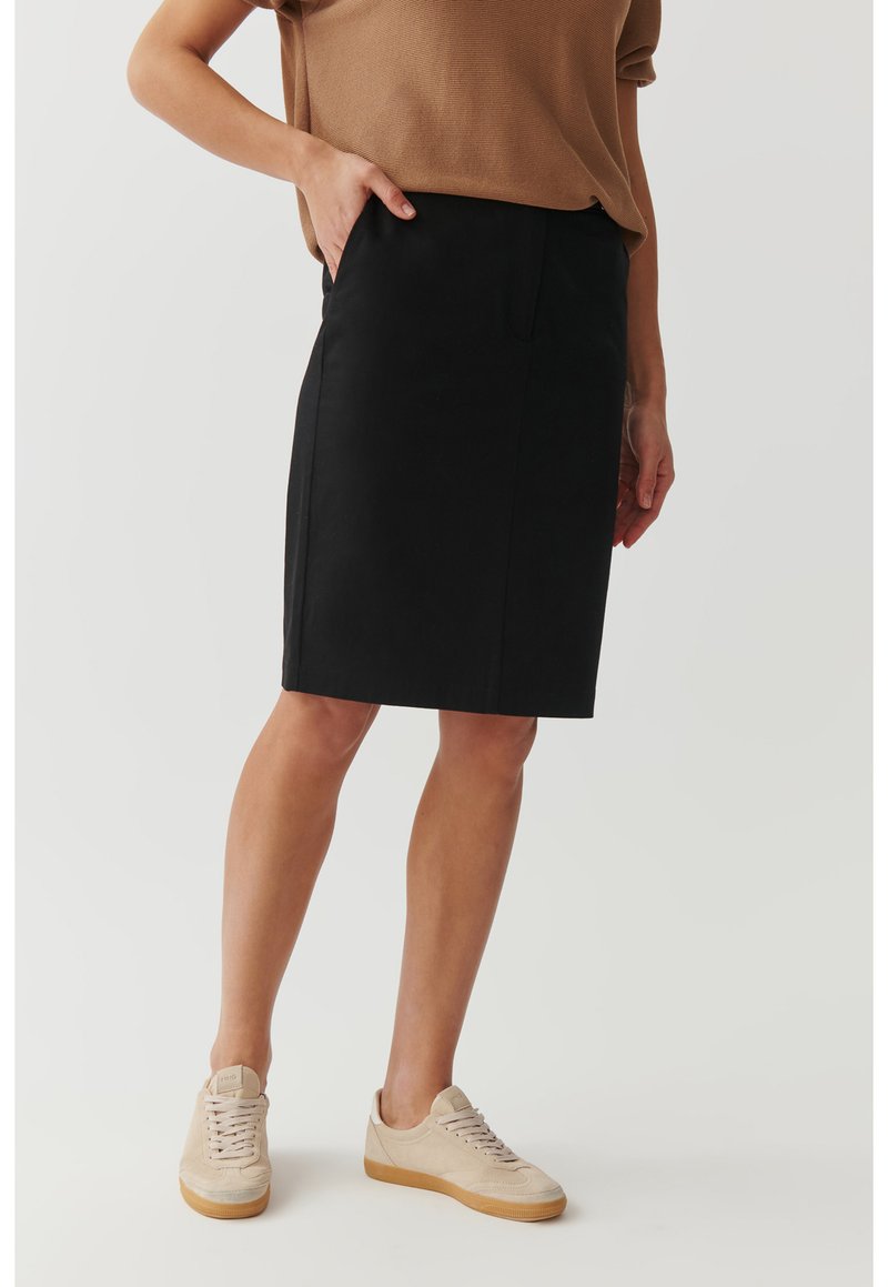 Čierna sukňa po kolená s elegantným dizajnom, vyrobená z hladkej tkaniny. Obsahuje bočné vrecká a je skombinovaná so svetlohnedými neformálnymi teniskami.