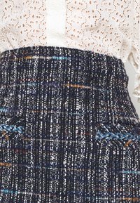 Gonna in tweed blu navy con sfumature multicolori, lavorata, con cintura spessa e tasche frontali. Blusa bianca ricamata con colletto a bottoni.