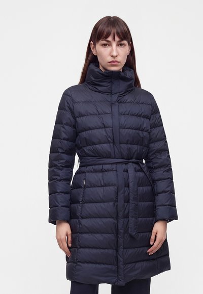 Femme portant un long manteau matelassé bleu marine avec un col montant et une ceinture nouée, debout devant un fond clair uni.