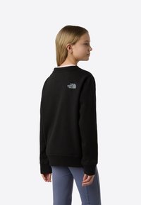 Μαύρο φούτερ με στρογγυλή λαιμόκοψη και ριμπ τελειώματα στα μανίκια, με μικρό γκρι λογότυπο στο πίσω μέρος. Φορεμένο με ανοιχτό μπλε παντελόνι.