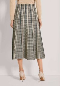 Frau in einem beigefarbenen und schwarzen karierten Midikleid mit vertikalen Streifen, beigem Pullover und beigefarbenen High Heels, die sich von der Kamera abwendet.