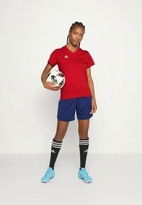 Piros V-nyakú mez, kék rövidnadrág, fekete térd zoknik fehér csíkokkal, egy fekete-fehér focilabdát tart, világoskék stoplis cipőt visel.