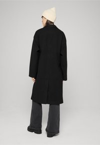 Melns ceļa garuma mētelis ar taisnu griezumu, sānu kabatām un vienu aizmugurējo šķēlumu, kopā ar pelēnām platajām biksēm un krēmkrāsas cepuri.