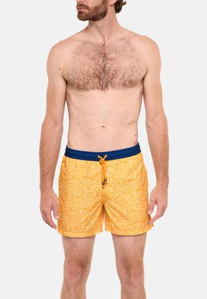 Homme torse nu avec poils sur la poitrine, portant un short de bain jaune à motif floral avec une ceinture bleu marine et un cordon, debout devant un fond uni.