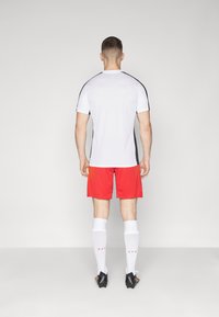 Korte mouwen witte sportshirt met zwarte accenten, gecombineerd met rode shorts, witte sokken en zwarte schoenen, met een sportief ontwerp.