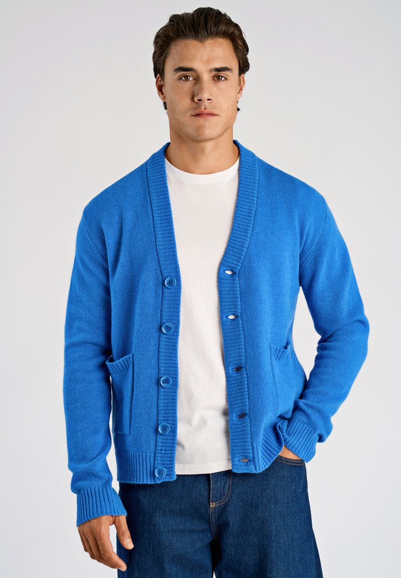 Blaue Strickjacke mit V-Ausschnitt, Vorderknöpfen und Taschen. Gerippte Bündchen und Saum. Wird über einem weißen T-Shirt getragen, kombiniert mit dunkelblauer Jeans.