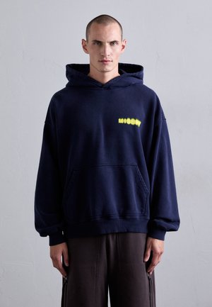 Man wearing navy blue hoodie with yellow "MISBHV" text, standing against plain light gray wall, hands relaxed by sides.