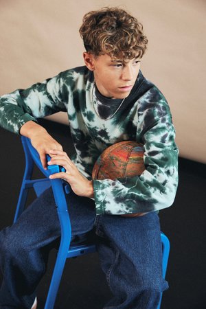 Felpa con un motivo tie-dye verde e nero, abbinata a pantaloni in denim blu. Tiene in mano un pallone da basket marrone con texture, seduto su una sedia blu.