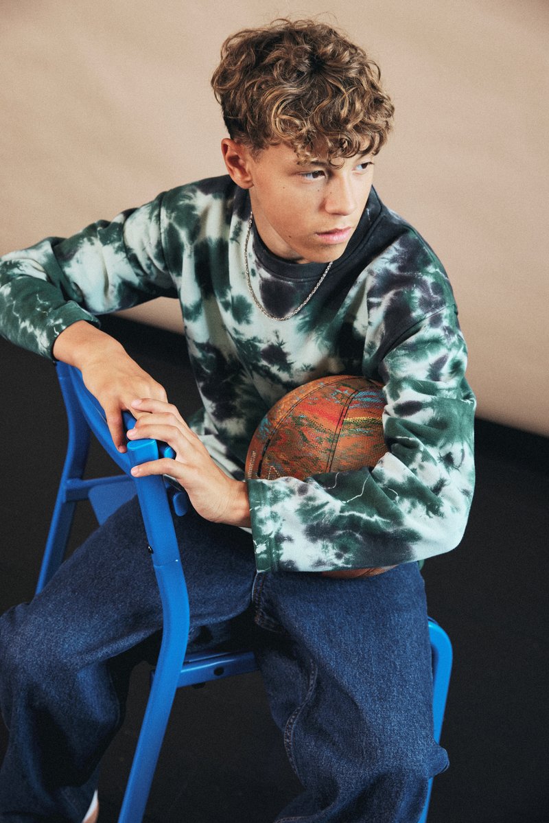 Sweatshirt with a tie-dye pattern in green and black, paired with blue denim pants. Holding a textured brown basketball, seated on a blue chair.