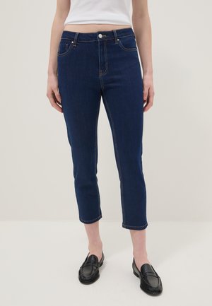 Femme portant un jean slim court bleu foncé, des mocassins noirs et un débardeur blanc court, debout devant un fond clair uni.