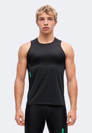 Jeune homme aux cheveux blonds courts portant un haut de sport noir sans manches et un pantalon noir assorti sur un fond clair uni.