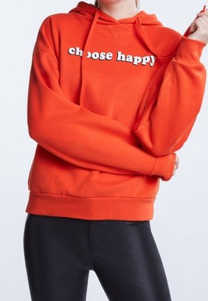 Osoba ubrana w jasnopomarańczową bluzę z napisem „choose happy” w białym kolorze, w połączeniu z czarnymi spodniami, na jednolitym tle.