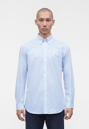 Homme portant une chemise à carreaux bleu clair avec col, debout les bras détendus sur fond blanc.