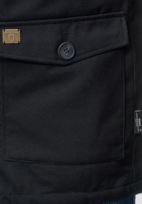 Poche en tissu texturé noir avec une fermeture à bouton et une étiquette en métal de marque. Caractérisée par un rabat proéminent et une étiquette latérale supplémentaire.