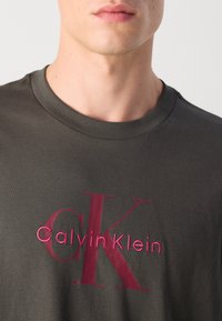 Tamno siva pamučna majica s velikim burgundskim CK logom ispod kojeg se nalazi tekst "Calvin Klein", rebrasti okrugli izrez i regularni kroj.