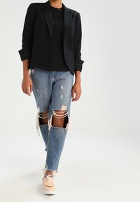 Blazer nero su una maglietta nera, abbinato a jeans chiari strappati con grandi strappi alle ginocchia. Le calzature includono mocassini beige.