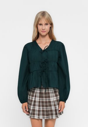 Jonge vrouw in een donkergroene blouse met lange mouwen en voorbinders, samen met een bruine en witte geruite plissé rok, staand tegen een effen achtergrond.
