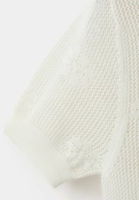 Gros plan sur un tissu tricot blanc avec des motifs floraux texturés et une bordure côtelée à l'extrémité de la manche.