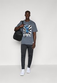 Pelēka grafiskā t-kreklā ar zilu tēlu un tekstu "SWOOSH", apvienots ar tumšiem biksēm un baltām kedām; tur melnu somu.