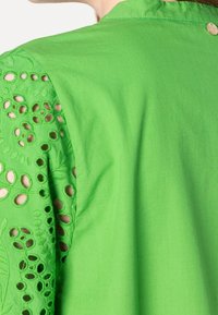 Blusa verde con mangas de encaje bordado de ojo detallado y espalda de tela lisa, cierre de botón dorado en el cuello.