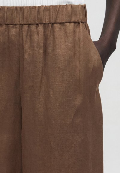 Καφέ λινό παντελόνι με ελαστική μέση που φοράει ένα άτομο με σκουρόχρωμο δέρμα, το χέρι ακουμπά στην δεξιά τσέπη.