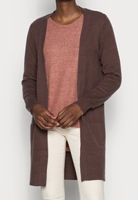 Brun cardigan med åben front, lange ærmer og to frontlommer over en kortærmet, lys pink, tekstureret top, som er parret med cremede bukser.