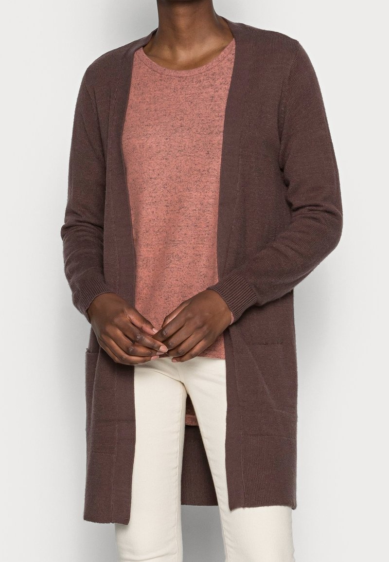 Brun cardigan med åben front, lange ærmer og to frontlommer over en kortærmet, lys pink, tekstureret top, som er parret med cremede bukser.