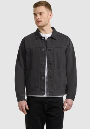 Homme portant une veste noire à boutons par-dessus un T-shirt blanc, debout devant un fond gris clair uni, regardant vers sa gauche.