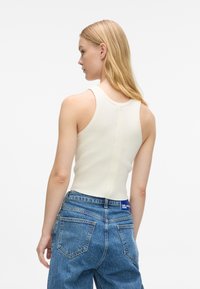 Biely rebrovaný tielko so priliehavým strihom, skombinované s vysoko pásovými modrými džínsami, ktoré majú štítok na páske a klasické vrecká.