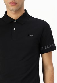 Čierna polo košeľa z bavlny s tradičným golierom. Obsahuje malé logo "GUESS" na hrudi a detail loga na rukáve.
