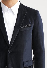 Blazer in velluto navy con revers a punta, chiusura a singolo bottone e taschino sul petto con inserto nero. Tessuto testurizzato.