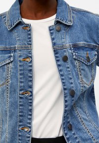 Giacca di denim di colore azzurro chiaro, con due tasche sul petto, bottoni scuri, camicia bianca sotto e cuciture arancioni a contrasto.
