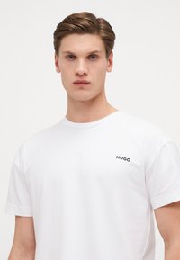 T-shirt en coton blanc avec un col rond et des manches courtes, ornée d'un petit logo noir "HUGO" sur la partie gauche de la poitrine.