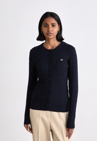 Marineblauer Cardigan mit Kabelstrickdesign, langen Ärmeln, Rundhalsausschnitt und Knöpfen an der Vorderseite. Kleines Logo-Detail auf der Brust.