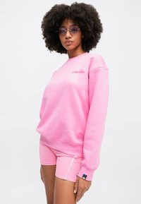 Ružový set mikiny a šortiek, vyrobený z jemného materiálu, s rebrovanými manžetami a detailom loga na hrudi. Hladká textúra a ležérny dizajn.