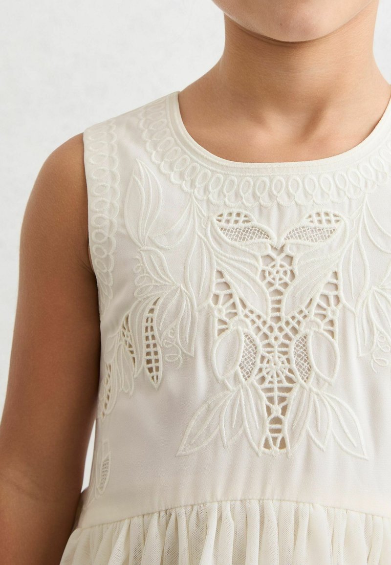 Kremna obleka z zapletenim belim vezenjem cvetličnih motivov na korzetu in nabranim tila krilom na spodnjem robu.