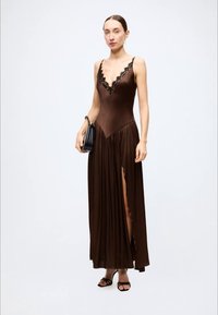 Brown satin evening dress with lace trim, deep v-neckline, and pleated skirt featuring a thigh-high slit. Model holds a small black bag.