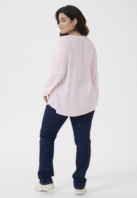 Camisa de manga larga rosa claro con un patrón texturizado, dobladillo curvado, combinada con jeans azul oscuro y zapatillas blancas.