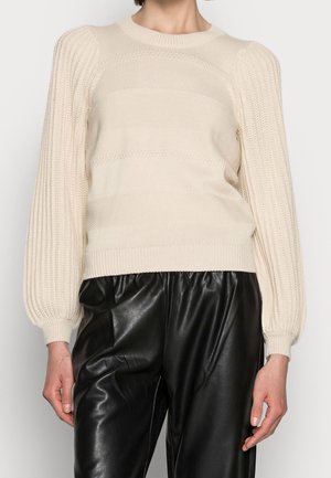 Femme portant un pull en tricot crème avec des manches bouffantes et un pantalon en cuir noir taille haute, debout devant un fond uni.